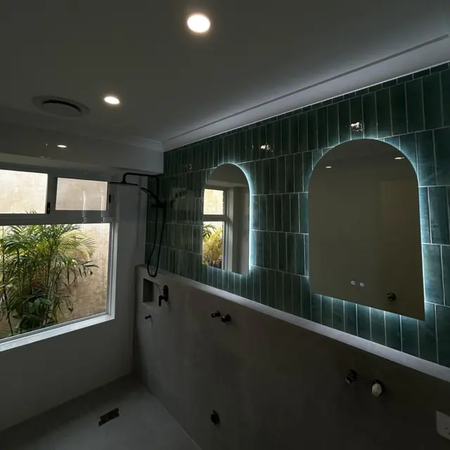 Two arched backlit LED mirrors on a green tiled bathroom wall
